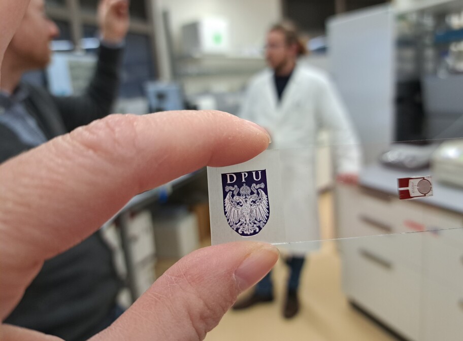 Eine Hand hält ein Muster eines Sensors mit dem Logo der DPU im Hintergrund ist...