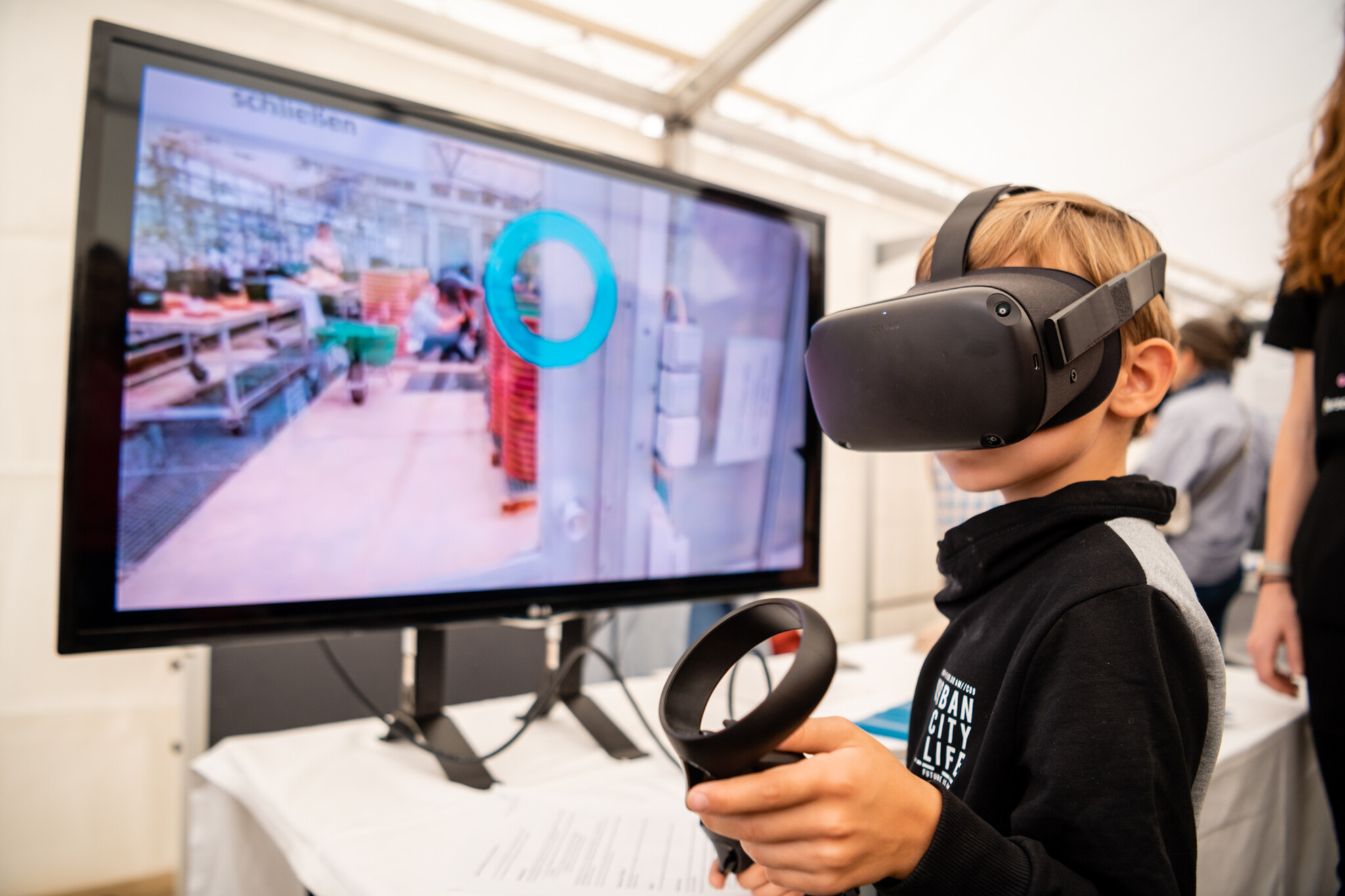 Kind hat VR Brille auf und im Hintergrund ist ein großer Bildschirm zu sehen