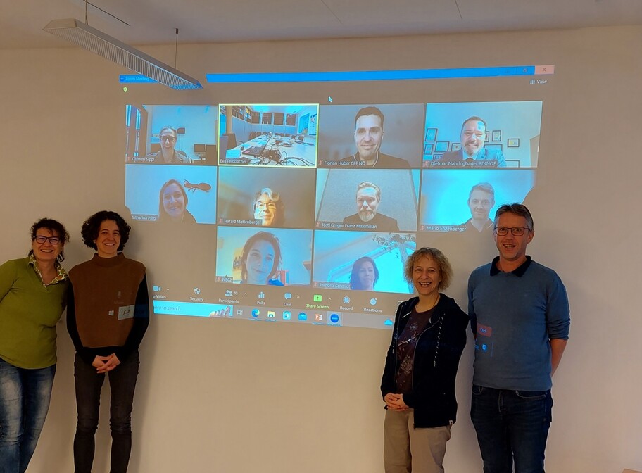 Kooperationsteilnehmer stehen vor dem Screen auf dem die online Teilnehmer zu...